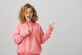 Come inside, this party is awesome. Studio shot of attractive curly-haired caucasian girl in eyewear and pink hoodie