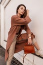 Comely attractive young woman in fashion beautiful elegant coat in beige pants stylish shoes with leather handbag rests near wall Royalty Free Stock Photo