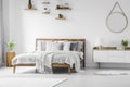 Comfortable big wooden framed bed with linen, pillows and blanket, nightstand beside and round mirror hanging on a white wall in