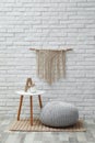 Comfortable knitted pouf, table and decor elements near white brick wall indoors. Interior design