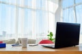 Comfortable workplace with laptop computer, supplies and cup of coffee, copy space. Wood table in sunny office with big windows.