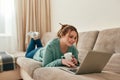 Comfortable workplace. Young beautiful girl in casual clothes lying on sofa in the living room, using laptop and Royalty Free Stock Photo