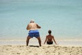 Comic and funny situation. An elderly man takes photos of back view of beautiful girl sitting on the beach