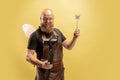 Comic portrait of muscular bearded bald man, blacksmith in leather apron or uniform isolated on yellow studio background