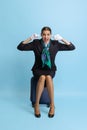 Comic portrait of pretty young girl, flight attendant, stewardess sitting on suitcase isolated on blue studio background