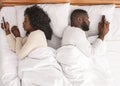 Afro couple using smartphones in bed, ignoring each other after quarrel Royalty Free Stock Photo