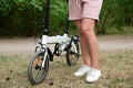 Compact folding bike next to the legs of an adult woman. A woman is standing in beige shorts and white sneakers