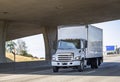 Compact medium power rig semi truck with box trailer running on the highway road under the bridge Royalty Free Stock Photo