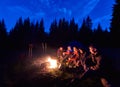 Company young people is sitting around fire against the backdrop of silhouetted large spruce trees and night sky Royalty Free Stock Photo