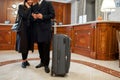 Comparing. Cropped shot of a couple in casual wear standing in hotel lobby with suitcase. Man is using his mobile phone Royalty Free Stock Photo