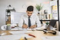 Competent arhitect indian male sitting on desk indoors using pen and ruler for project drawings.