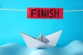 Competition concept. Paper boat among waves and red sign with word Finish on light blue background Royalty Free Stock Photo