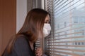 Compliance with quarantine. A young woman in an antibacterial mask looks out the window from the house to the street, sits in the