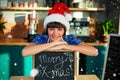 Composite image of portrait of smiling waitress wearing santa hat and sitting with x-mas sign board Royalty Free Stock Photo