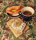 Composition of banana puff on a plate, a blue cup of tea, a fork, two notebooks and a citrine stone on a multi-colored tablecloth. Royalty Free Stock Photo