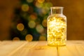 Composition of fairy lights in vase on wooden table with christmas tree blurred in background Royalty Free Stock Photo