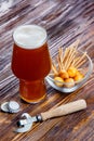 Composition of a glass of beer with foam on a rustic wooden table next to a bowl of salty snacks and a bottle opener