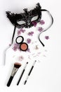 A composition of a masquerade black mask, spring flowers and cosmetics on a white background. Purple flowers are scattered