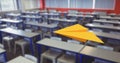 Composition of yellow paper aeroplane flying over desks in empty classroom