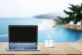 Computer laptop with black screen and hot coffee cup on wooden table top on blurred pool and beach background Royalty Free Stock Photo