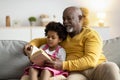 Concentrated african american small curly girl and smiling elderly man reading book with fairy tales Royalty Free Stock Photo