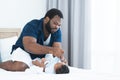 Concentrated African Nigerian bearded father changing diaper and clothes for newborn baby lying on white bed in bedroom. Single
