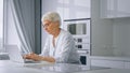 Concentrated aged businesswoman with short grey hair and glasses types on laptop sitting in kitchen at home