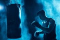 Concentrated boxer in boxing gloves training with punching bag in dark.