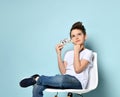 Concentrated schoolboy sits on chair holding dollars and touching chin with hand thinking how to earn more.