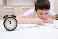 A concentrated boy writing in a textbook, exercisebook, a schoolboy doing his homework at home, an alarm clock in front Royalty Free Stock Photo