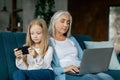 Concentrated busy caucasian small girl plays on smartphone, old lady works on laptop on sofa
