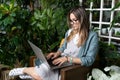 Concentrated female freelancer sitting on chair in green house, working on laptop surrounded by plants. Remote work Royalty Free Stock Photo