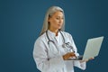 Concentrated grey haired doctor works on laptop standing on blue background