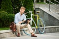 Concentrated, handsome young businessman sitting on bench outside office with bike and working online on tablet Royalty Free Stock Photo