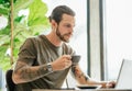Concentrated hipster man with bearded working on laptop and drinking coffee or tea at coffee shop. Online marketing, education, e- Royalty Free Stock Photo