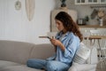 Concentrated introverted woman with smartphone sit on sofa with legs tucked under in living room