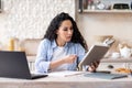 Concentrated lady sitting in kitchen, using laptop and reading book, studying remotely from home