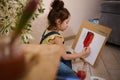 Adorable concentrated little Caucasian girl in yellow t-shirt and blue denim drawing picture at home, using watercolor paints, Royalty Free Stock Photo