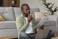 Concentrated pensive middle aged african american guy in glasses with laptop reads document Royalty Free Stock Photo