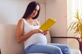 Concentrated pregnant woman reading book while sitting on sofa