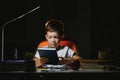 Concentrated schoolboy reading book at table with books, plant, lamp, colour pencils, apple, and textbook