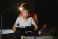 Concentrated schoolboy reading book at table with books, plant, lamp, colour pencils, apple, and textbook