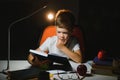 Concentrated schoolboy reading book at table with books, plant, lamp, colour pencils, apple, and textbook