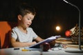 Concentrated schoolboy reading book at table with books, plant, lamp, colour pencils, apple, and textbook Royalty Free Stock Photo