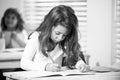 Concentrated schoolgirl sitting at desk and writing in exercise book with classmate sitting behind. Royalty Free Stock Photo