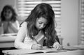 Concentrated schoolgirl sitting at desk and writing in exercise book with classmate sitting behind. Royalty Free Stock Photo