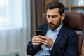 Concentrated and serious young businessman sitting in a chair in the office at the table and using a mobile phone Royalty Free Stock Photo