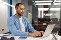 Concentrated and serious young hispanic man working in a modern office, sitting at a desk and typing on a laptop Royalty Free Stock Photo