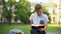 Concentrated teenager reading book in park on bench, preparing for exams
