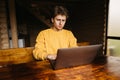 Concentrated young freelancer man in orange sweater sitting at table in room with wooden interior and working on laptop Royalty Free Stock Photo